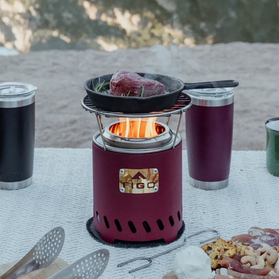 Portable camping fire pit with food cooking outdoors, surrounded by camping utensils and drinks.