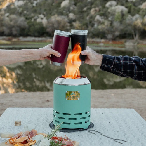 Two people toasting with flamed drinks over a portable fire pit by a lake.