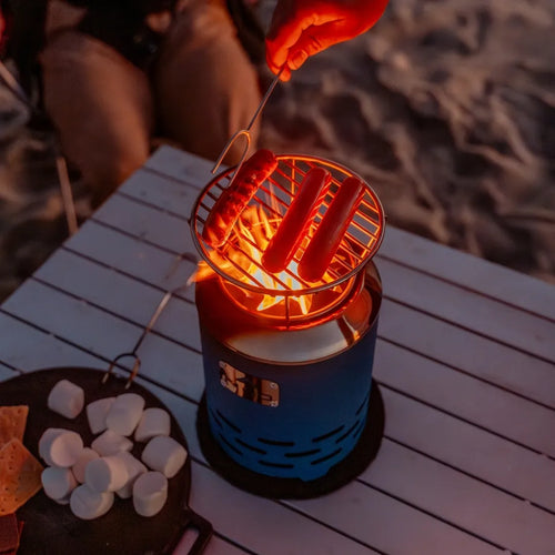 Person grilling sausages over a portable fire pit with grill, on a wooden table outdoors.